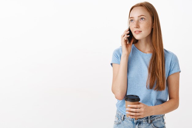 Mujer guapa de ensueño con mirada relajada sosteniendo una taza de café de papel mientras habla por teléfono