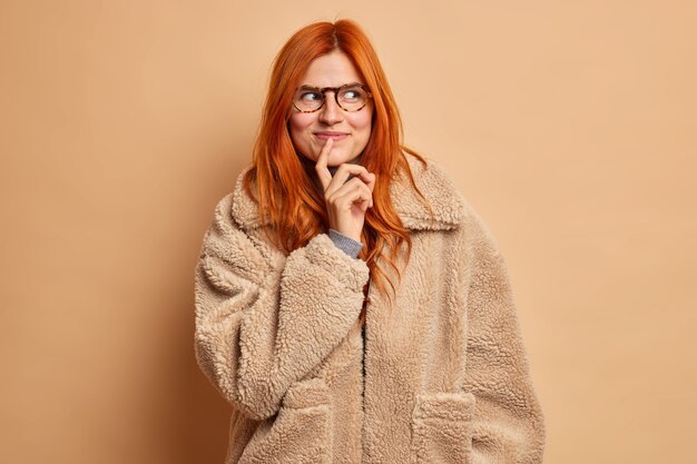 Una mujer guapa de ensueño con el pelo pelirrojo mira a un lado pensativamente y viste un abrigo de invierno marrón.