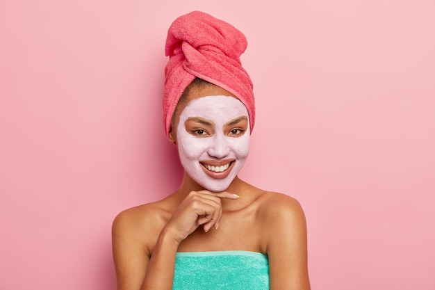 Mujer guapa con expresión complacida, toca la barbilla suavemente, usa mascarilla de arcilla limpiadora en la cara, tiene una toalla envuelta en la cabeza, disfruta de tratamientos de belleza en casa, aislada en la pared rosa