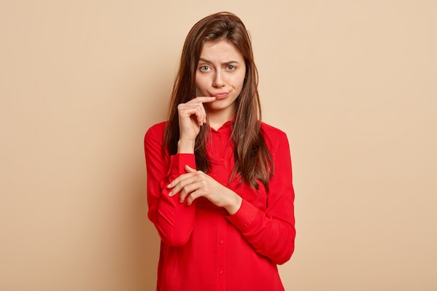 Una mujer guapa insatisfecha levanta las cejas, frunce los labios, mira con enojo, mantiene la mano cerca de la boca, usa una camisa roja elegante, se para sobre una pared beige, descontento con la sugerencia de alguien