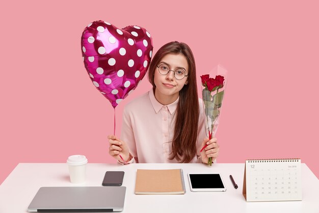 Foto gratuita una mujer guapa tiene una mirada atenta, recibe agradables regalos de su novio en la oficina, sostiene un globo de san valentín y rosas, usa gafas