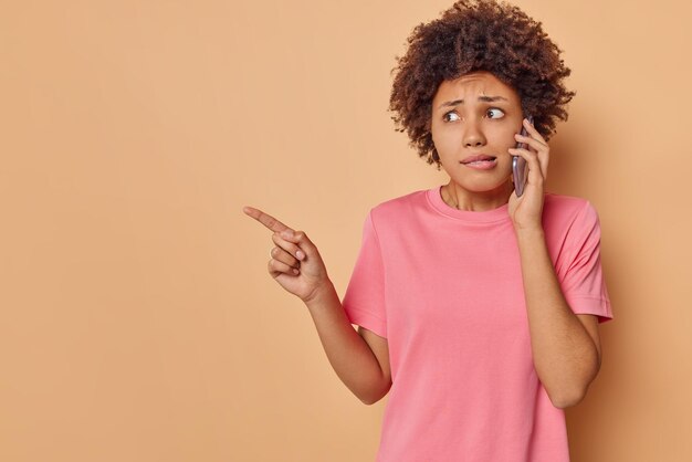 la mujer habla a través de un teléfono inteligente indica que se siente asustada y tiembla de miedo muestra que el espacio de la copia en blanco usa una camiseta rosa aislada en beige. Oh, no mires ahí