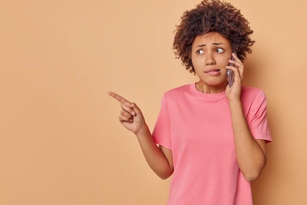 la mujer habla a través de un teléfono inteligente indica que se siente asustada y tiembla de miedo muestra que el espacio de la copia en blanco usa una camiseta rosa aislada en beige. Oh, no mires ahí