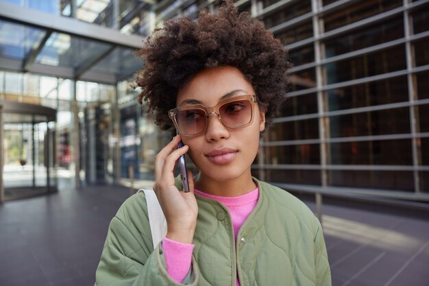 La mujer habla a través del teléfono móvil utiliza una buena conexión en itinerancia disfruta de un recorrido por la ciudad se comunica con un amigo lleva gafas de sol chaqueta plantea fuera