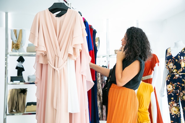 Mujer hablando por celular mientras elige ropa y busca vestidos en rack en tienda de moda. Plano medio, vista lateral. Cliente boutique o concepto minorista