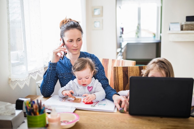 Mujer hablando por teléfono inteligente mientras sus hijos juegan en escritorio