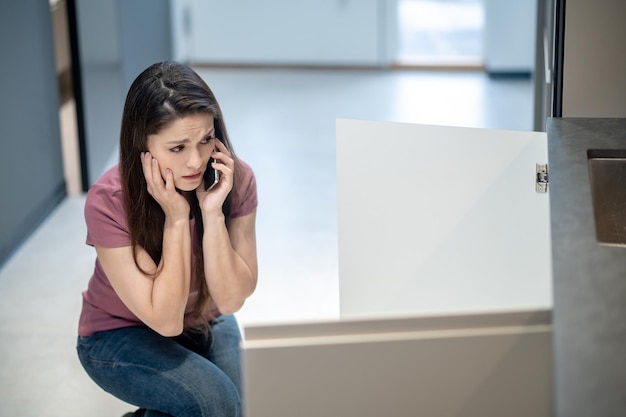 Mujer hablando por teléfono inteligente mirando debajo del fregadero