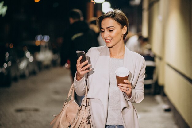 Mujer hablando por teléfono y tomando café afuera en la calle por la noche
