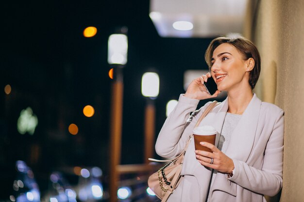 Mujer hablando por teléfono y tomando café afuera en la calle por la noche