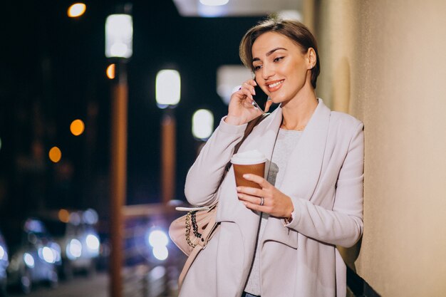 Mujer hablando por teléfono y tomando café afuera en la calle por la noche