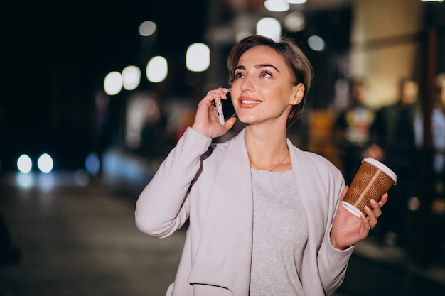 Mujer hablando por teléfono y tomando café afuera en la calle por la noche