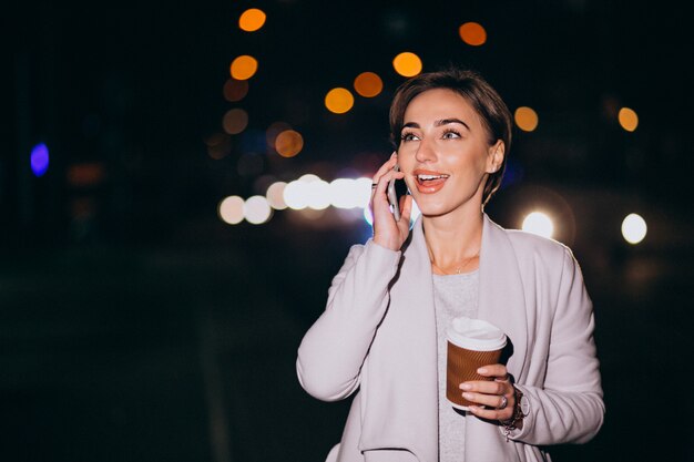 Mujer hablando por teléfono y tomando café afuera en la calle por la noche