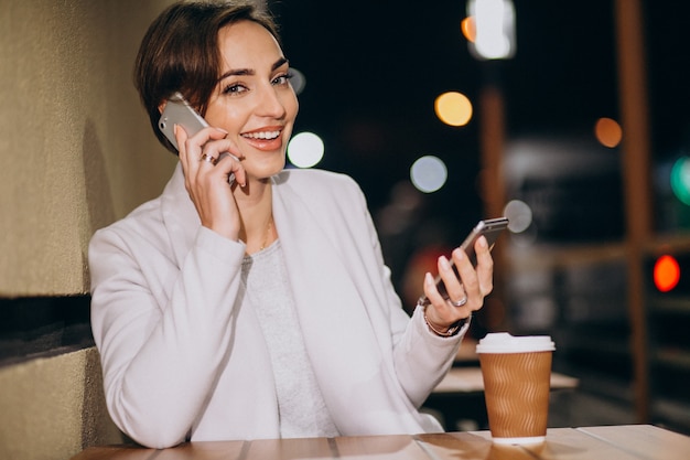 Mujer hablando por teléfono y tomando café afuera en la calle por la noche