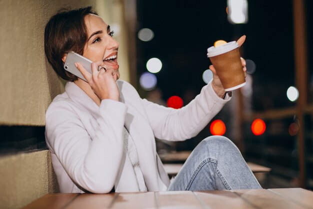 Mujer hablando por teléfono y tomando café afuera en la calle por la noche