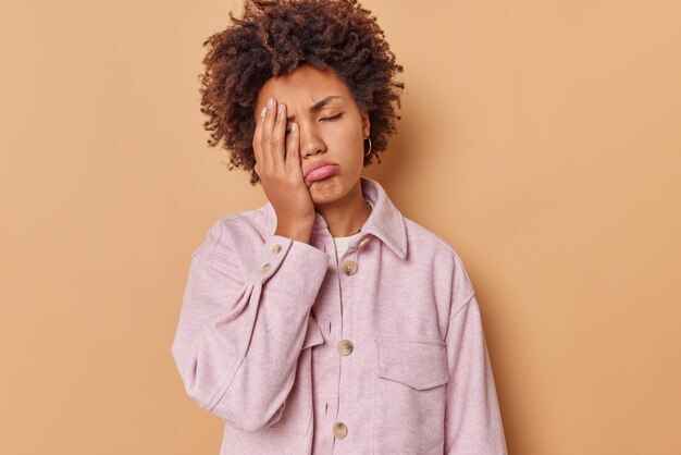 mujer hace cara palma tiene expresión sombría mantiene los ojos cerrados usa chaqueta rosa aislada en beige se siente cansada quiere dormir después de largas horas de trabajo