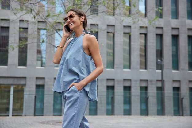 La mujer hace una conversación internacional con un teléfono inteligente, usa un traje de moda, gafas de sol, disfruta de llamadas de teléfono celular, camina al aire libre cerca del edificio de la ciudad moderna