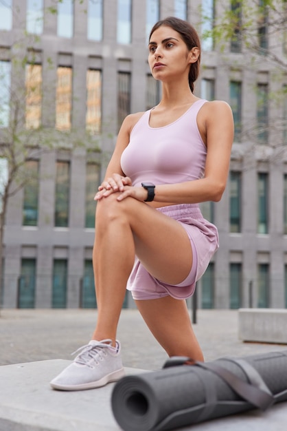 Foto gratuita mujer hace ejercicio al aire libre vestida con traje deportivo lleva zapatillas plantea en la ciudad utiliza karemat para entrenamiento físico