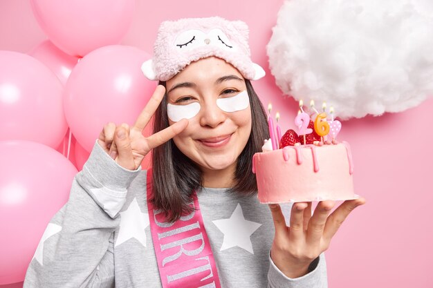 Mujer hace gesto de paz en los ojos sonríe agradablemente tiene un estado de ánimo alegre sostiene un sabroso pastel celebra cumpleaños