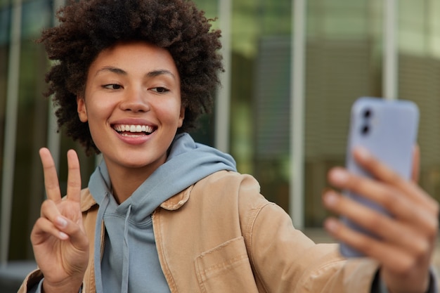 Mujer hace gesto de paz toma selfie en la cámara del teléfono inteligente hace llamadas en línea vestida con ropa casual plantea al aire libre