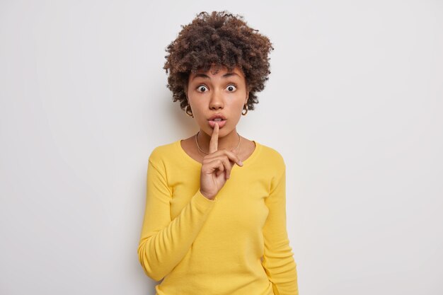 Mujer hace gesto de silencio mantiene el dedo índice en los labios chismes sobre algo vestido con un jersey amarillo casual sobre blanco