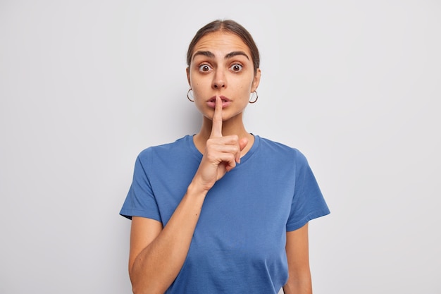 mujer hace gesto de silencio presiona el dedo índice en los labios vestida con una camiseta azul casual pide que se mantenga callado hace un signo tabú posa en blanco chismes sobre algo
