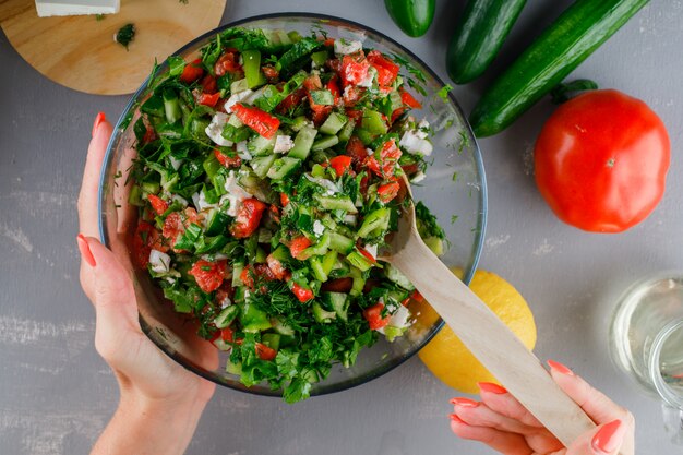 Mujer haciendo ensalada de verduras en un recipiente de vidrio con tomates en una vista superior de la superficie gris