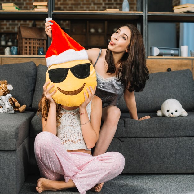 Mujer haciendo expresión divertida mientras sostiene emoticon de santa claus smiley frente a la cara de su amiga