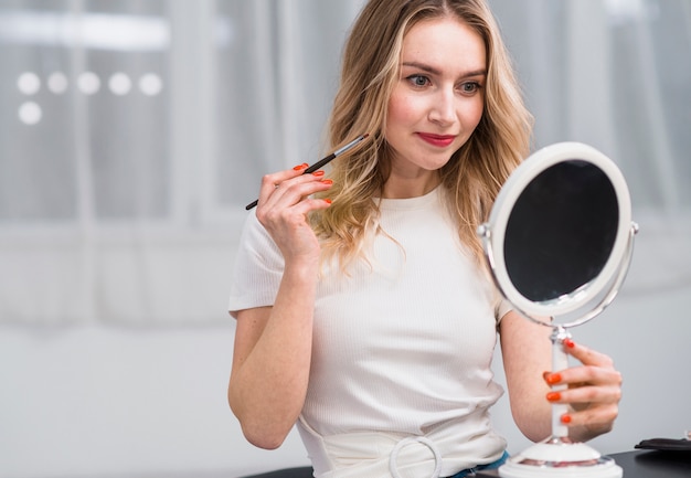 Mujer haciendo maquillaje mientras sostiene el espejo