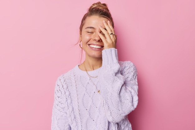 Una mujer hermosa y alegre hace que la palma de la cara cierre los ojos y sonríe alegremente con los dientes, tiene el cabello peinado, usa poses de puente de punto púrpura contra un fondo rosa. Concepto de personas y felicidad