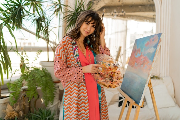 Mujer hermosa artista en traje bohemio posando con pincel y paleta en su estudio de arte.