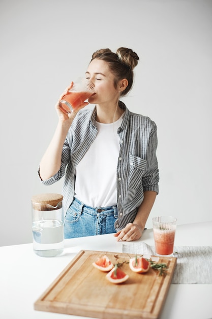 Mujer hermosa con los bollos que sonríen bebiendo el batido de desintoxicación del pomelo sobre la pared blanca. Dieta saludable nutrición