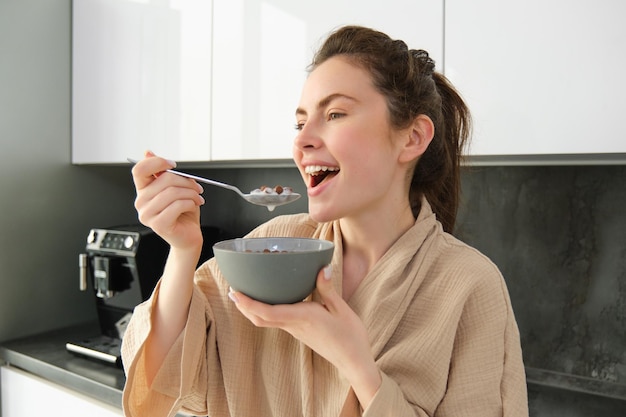 Una mujer hermosa comienza su día con cereales, desayuna y sonríe, usa una bata de baño y se apoya en la cocina