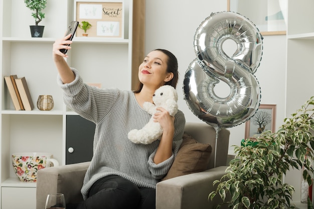 Mujer hermosa complacida en el día de la mujer feliz con oso de peluche tomar un selfie sentado en un sillón en la sala de estar