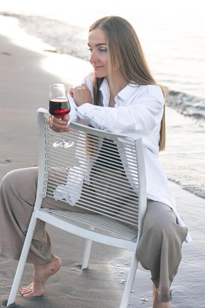 Una mujer hermosa con una copa de vino a la orilla del mar se sienta en una silla