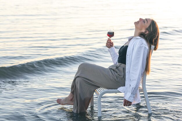 Una mujer hermosa con una copa de vino a la orilla del mar se sienta en una silla