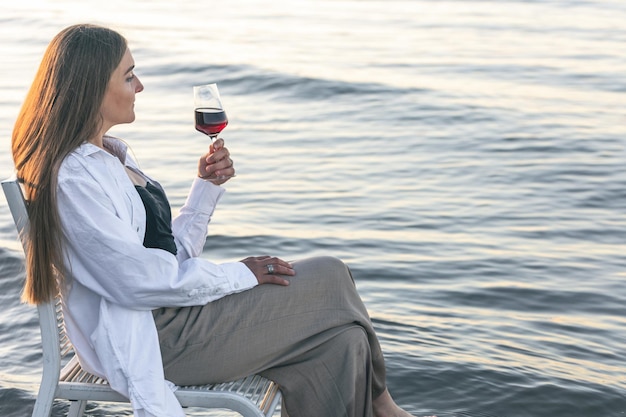 Foto gratuita una mujer hermosa con una copa de vino a la orilla del mar se sienta en una silla