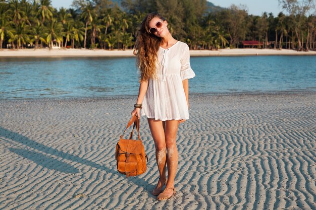 Mujer hermosa delgada en vestido de algodón blanco caminando en la playa tropical en la puesta del sol con mochila de cuero.