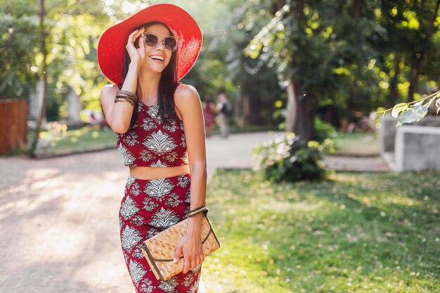 Mujer hermosa elegante caminando en el parque en traje tropical. dama en tendencia de moda de verano estilo callejero con bolso de paja, sombrero rojo, gafas de sol, accesorios. niña sonriendo feliz de vacaciones.