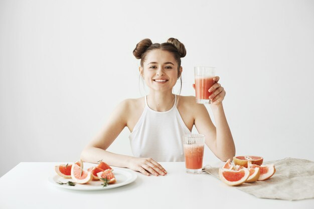 Mujer hermosa feliz que sonríe sentado en la mesa que se sostiene de cristal con el smoothie de pomelo fresco de desintoxicación sano sobre la pared blanca