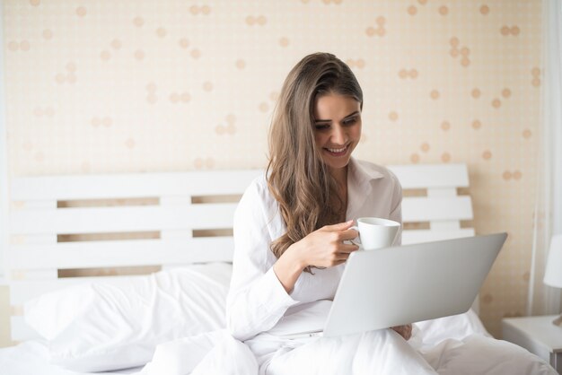 Mujer hermosa feliz que trabaja en una computadora portátil en la cama en la casa