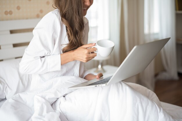 Mujer hermosa feliz que trabaja en una computadora portátil en la cama en la casa