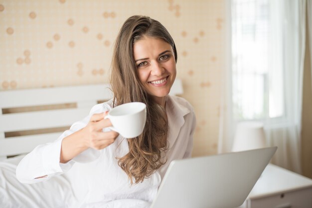 Mujer hermosa feliz que trabaja en una computadora portátil en la cama en la casa