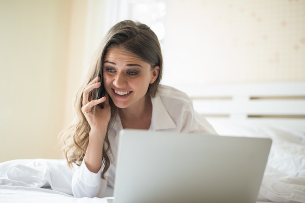 Mujer hermosa feliz que trabaja en una computadora portátil en la cama en la casa