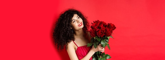 Foto gratuita mujer hermosa feliz en vestido sosteniendo flores y sonriendo romántico mirando a un lado en el logotipo de pie a