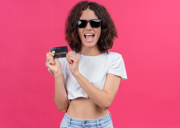 Mujer hermosa joven alegre con gafas de sol y sosteniendo la tarjeta en la pared rosa aislada con espacio de copia