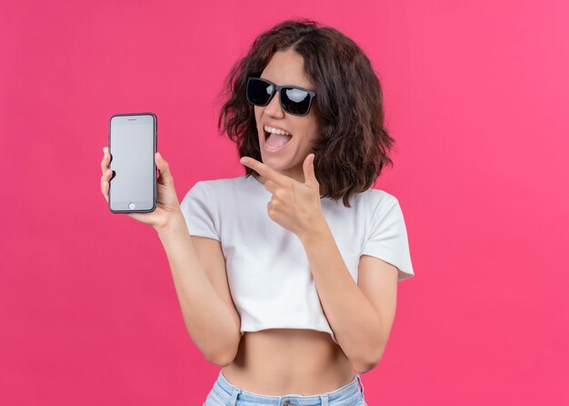 Mujer hermosa joven alegre con gafas de sol y sosteniendo el teléfono móvil apuntando a él en la pared rosa aislada con espacio de copia