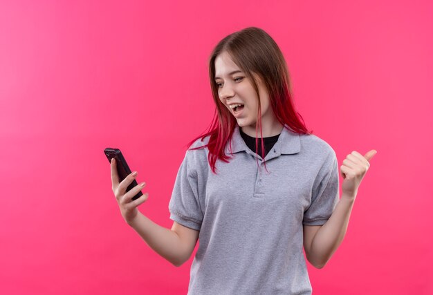 Mujer hermosa joven alegre que lleva la camiseta gris que mira el teléfono en la mano que muestra el gesto sí en la pared rosada aislada