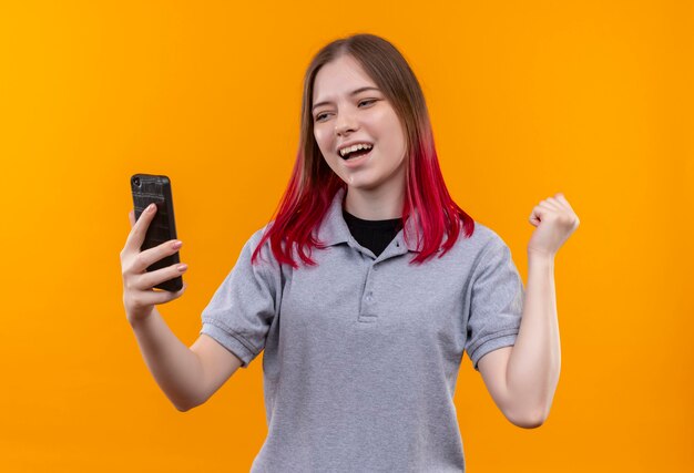 Mujer hermosa joven alegre que lleva la camiseta gris que mira el teléfono en su mano que muestra el gesto sí en la pared amarilla aislada