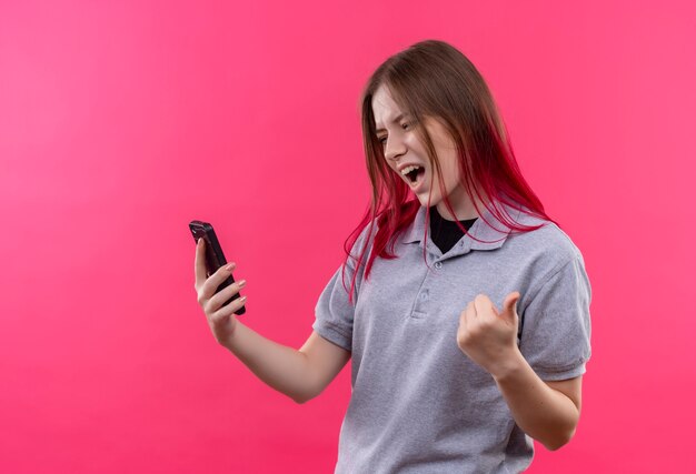 Mujer hermosa joven alegre que lleva la camiseta gris que mira el teléfono en su mano que muestra el gesto sí en la pared rosada aislada