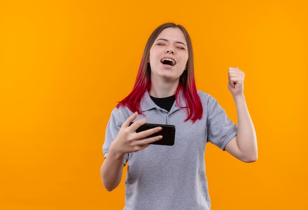 Mujer hermosa joven alegre que lleva la camiseta gris que sostiene el teléfono que muestra el gesto sí en la pared amarilla aislada